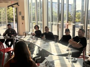 People sit around a table in a sun-soaked room. They are in discussion with paper around them.