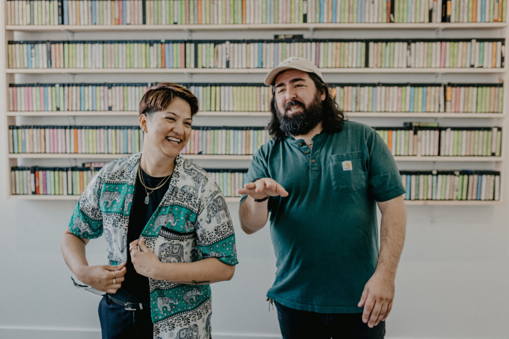 Two people look directly at the camera in front of a wall of VHS tapes 