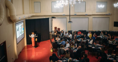 Salamishah Tillet, a Black woman in orange pants and a blue shirt, speaks in front of a crowd while gesturing to a screen.