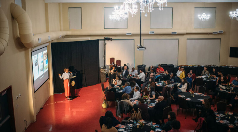 Salamishah Tillet, a Black woman in orange pants and a blue shirt, speaks in front of a crowd while gesturing to a screen.
